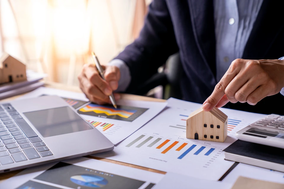 Scène d'intérieur bureau, homme en costume analysant des données immobilières, manipulant maquette maison en bois, stylo en main, consultant graphiques et tableaux de bord