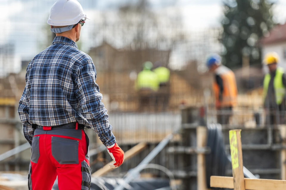 Chantier de construction intérieur avec trois professionnels du bâtiment en équipements de protection individuelle (casques jaune, bleu, blanc, gilets haute visibilité) analysant des plans architecturaux sur table de travail temporaire